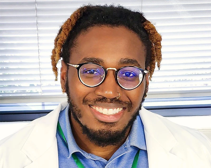 Dr. Ese smiling in a brightly lit office, wearing round glasses, a white lab coat, a blue collared shirt, and a green lanyard. His hair is styled in short twists with light brown tips, and he has a nose ring and neatly groomed beard.