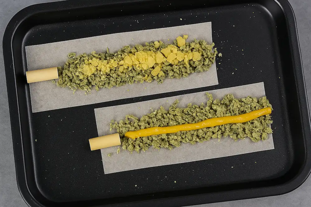 Two unrolled cannabis pre-rolls on parchment paper set on a black tray. The top one is filled with ground cannabis and yellow crumble concentrate, while the bottom one contains ground cannabis with a strip of golden cannabis oil or rosin down the center.
