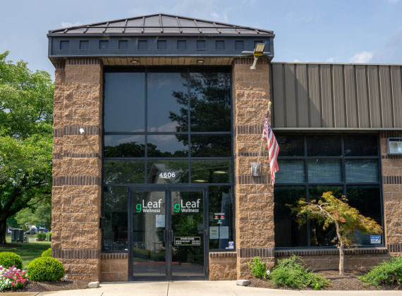 Exterior of a gLeaf Wellness dispensary in Frederick, featuring a modern yet natural architectural design. The building has a mix of brown brick and large glass windows, with a peaked metal roof over the main entrance. The entrance has double glass doors with the 'gLeaf Wellness' logo displayed on them. An American flag is mounted on the right side of the entrance, and the landscaping includes neatly trimmed bushes, flowers, and a small tree. The sky is partly cloudy, and the building is surrounded by a green suburban environment.