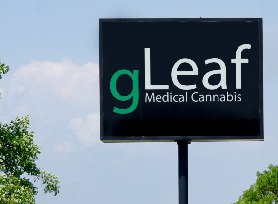 A gLeaf Medical Cannabis sign, featuring the brand's signature black background with "gLeaf" in bold white and green lettering, followed by "Medical Cannabis" in smaller white text. The sign is mounted on a tall pole, with a bright blue sky and some green tree branches visible in the background.